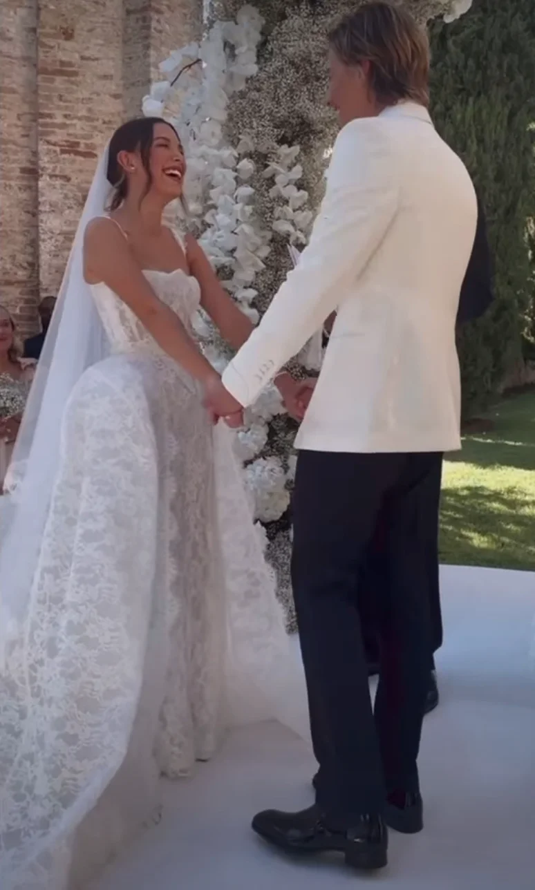 A couple holding hands at a wedding, smiling at each other. The bride wears a lace gown, and the groom is in a white jacket and dark trousers