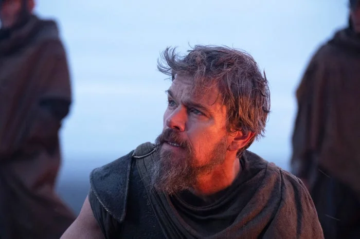 A man with a rugged beard and disheveled hair is wearing a tunic, appearing intense and focused in an outdoor setting with others in the background