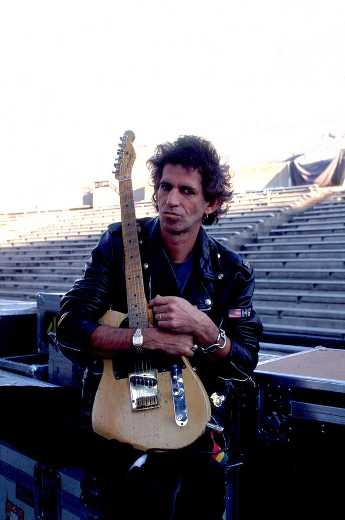 Keith Richards wears a leather jacket and holds a guitar while sitting on equipment cases at an outdoor venue
