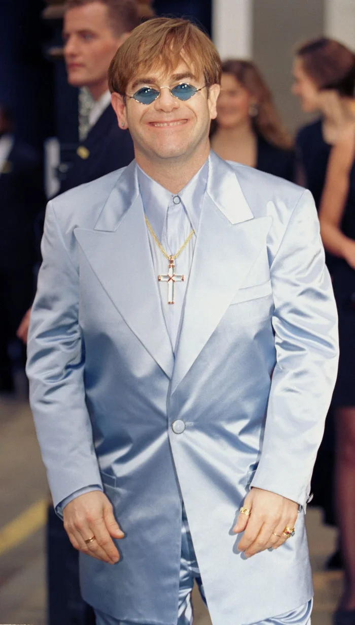 Elton John, in a shiny suit and round sunglasses, smiles while wearing layered gold necklaces