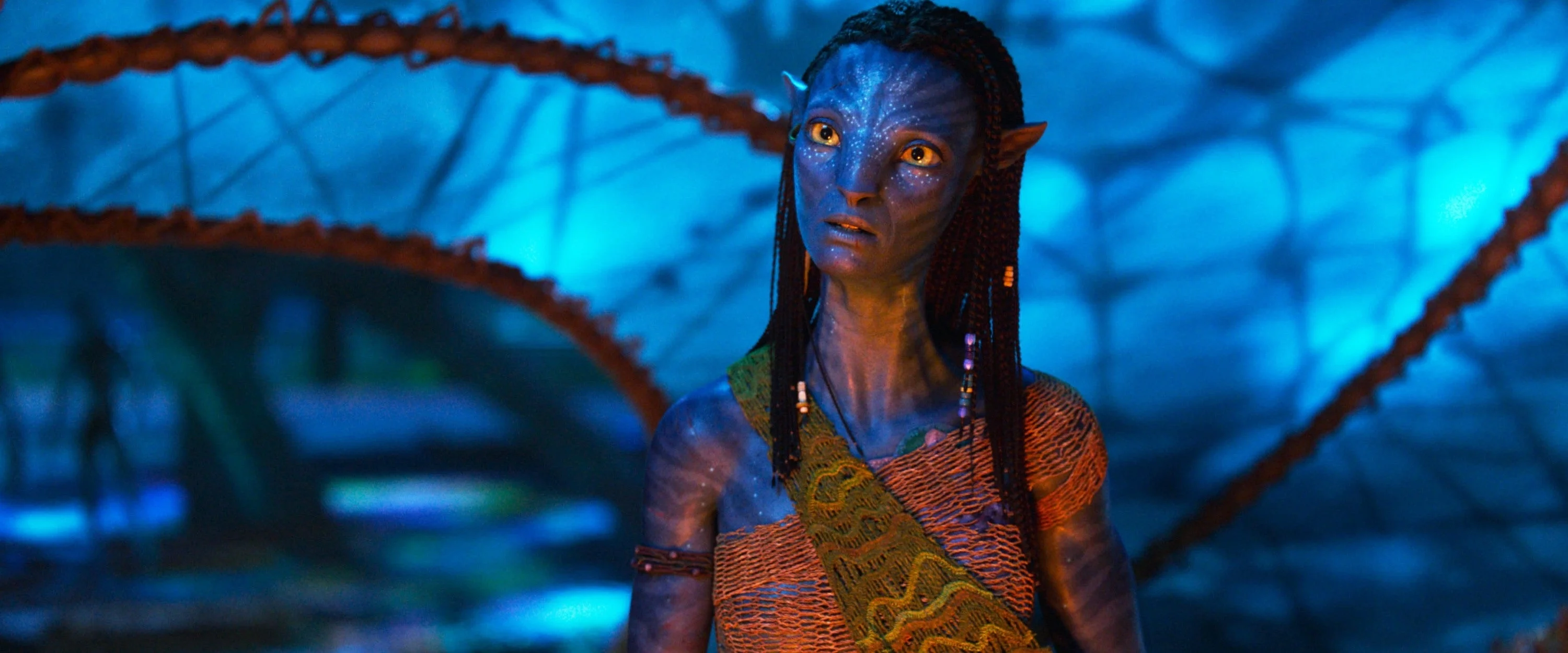 Zoe as a blue alien with pointed ears and braided hair stands in a glowing, bioluminescent forest wearing a patterned garment