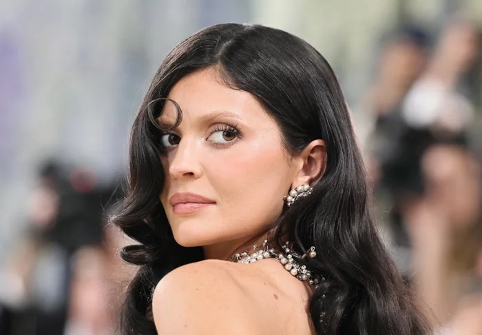 Kylie Jenner with long wavy hair, wearing a beaded necklace, looking over their shoulder at an event
