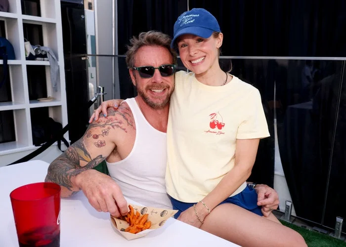 Man in tank top and sunglasses sits at a table with fries. Woman in casual tee and cap stands beside him, smiling. Casual outdoor setting