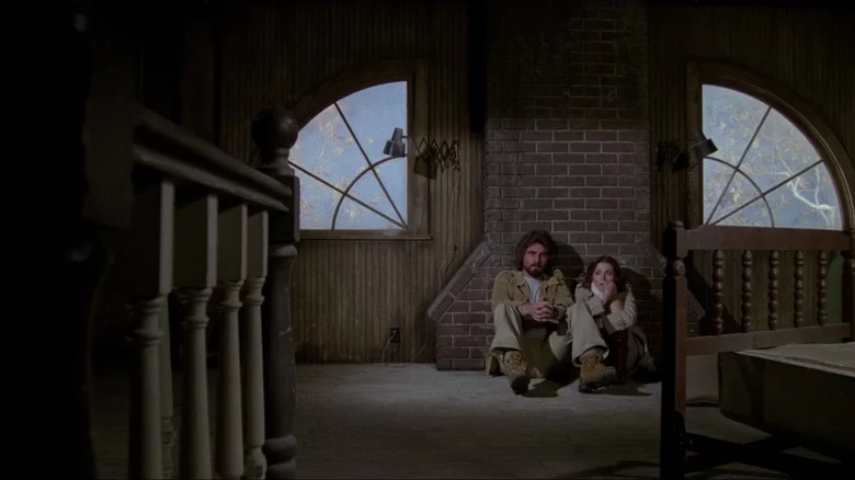 James Brolin as George Lutz and Margot Kidder as Kathy Lutz in The Amityville Horror, sitting on the ground next to each other against a brick wall in a dark, creepy-looking part of the house.