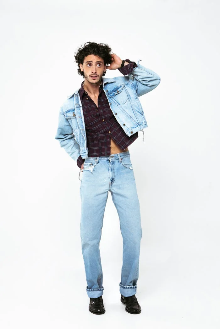 Person in denim jacket and jeans with a patterned shirt, striking a casual pose against a plain background