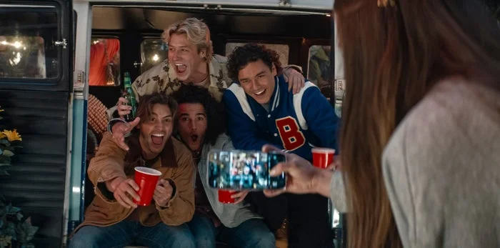 A group of four young people excitedly pose with drinks inside a van as someone takes their photo with a smartphone