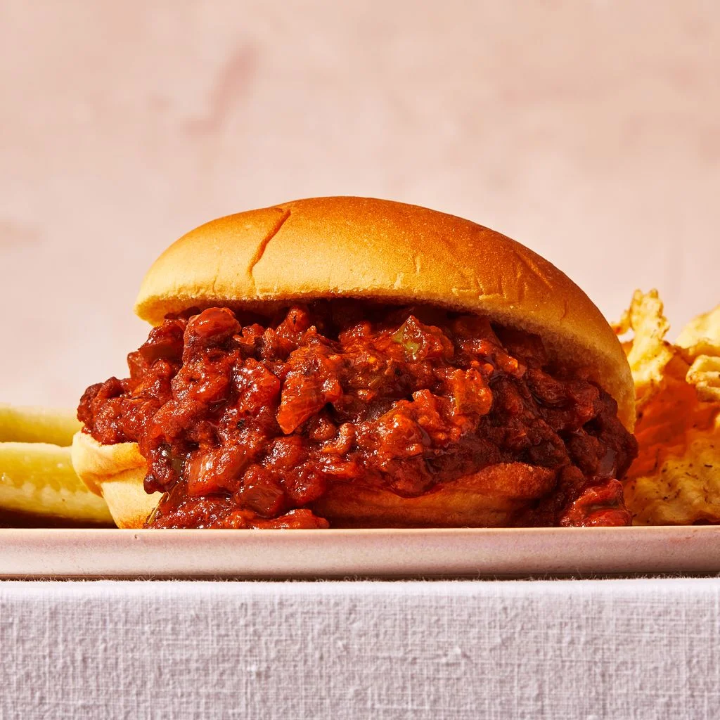 Closeup of a gochujang sloppy joe on a bun with ridged potato chips and pickles behind it