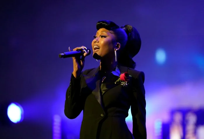 A person sings passionately on stage, wearing an elegant, dark suit with a red rose pinned to the lapel