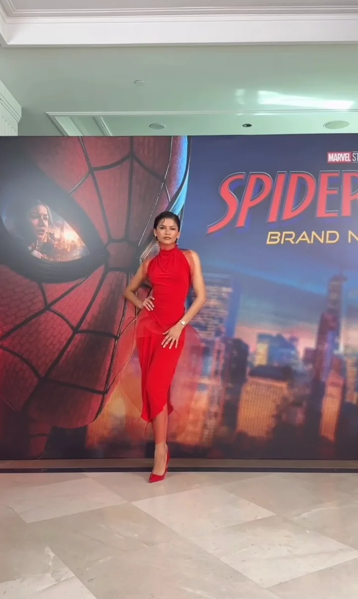 Zendaya poses in a sleek, fitted red dress and heels in front of a "Spider-Man: Brand New" poster at an event