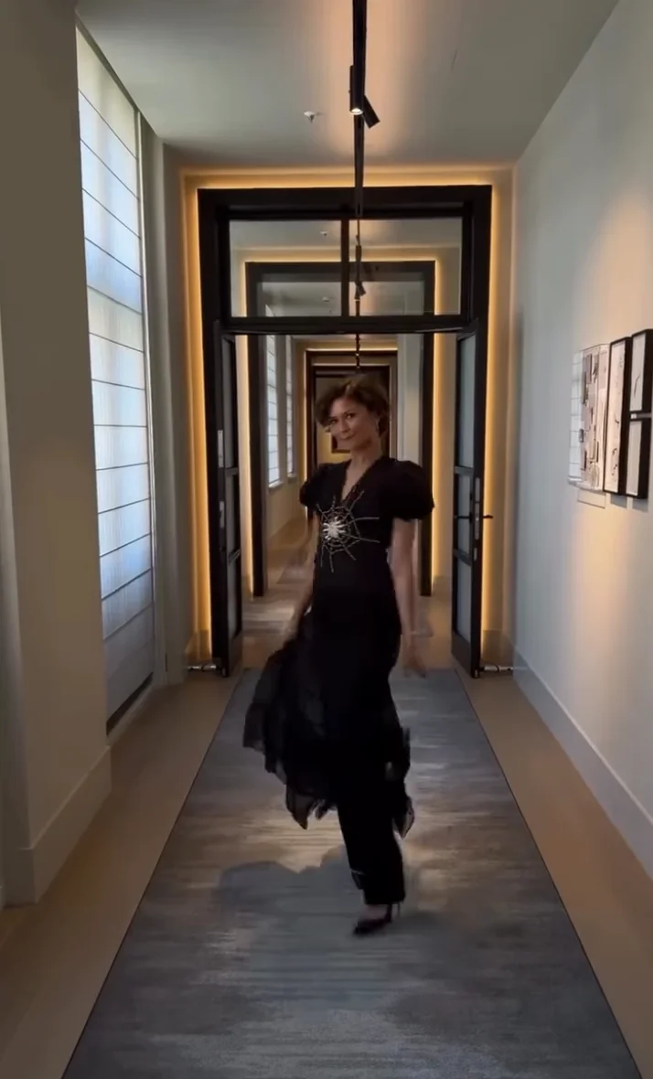 Zendaya walking down a hallway in a stylish black dress with puffed sleeves and a decorative design on the front