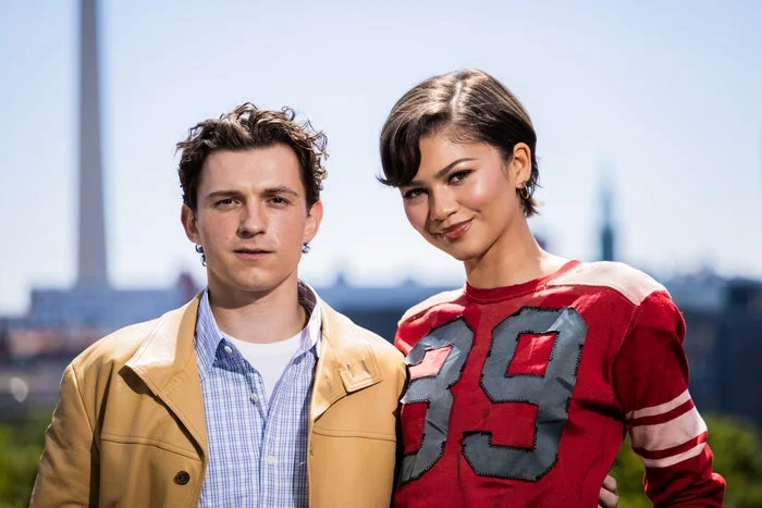 Zendaya and Tom standing outdoors. The person on the left wears a casual jacket and shirt. The person on the right wears a red top with "39" on it