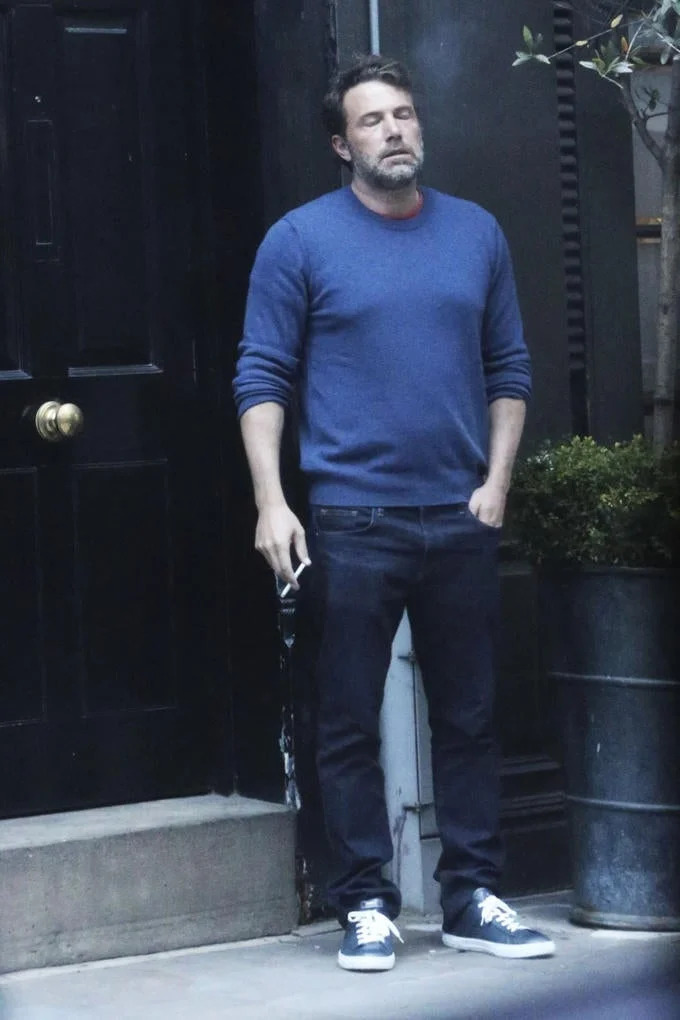 Ben Affleck standing outside a building while smoking a cigarette with an exhausted look and his eyes closed