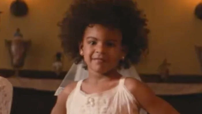 Child smiling confidently, wearing a sleeveless dress and a veil, standing indoors with warm lighting in the background