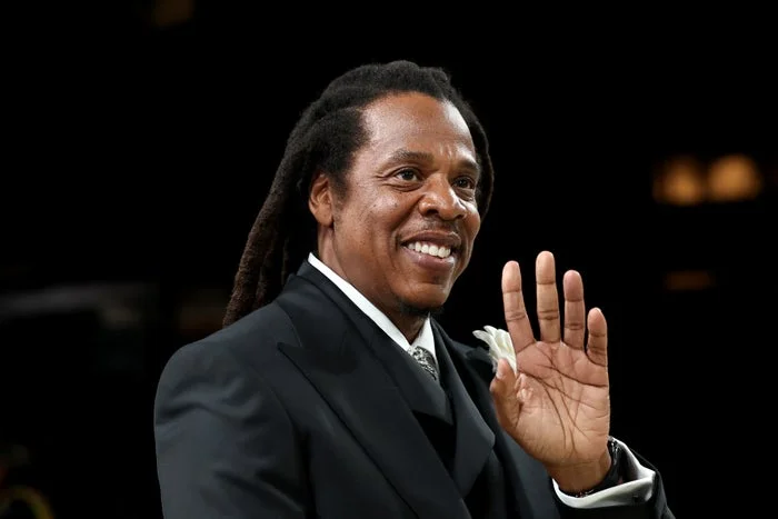 A person with long hair in a formal suit, smiling and waving, stands on a black background