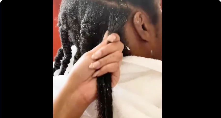 A person is applying hair product to sectioned braids on another person wearing a white towel over their shoulders
