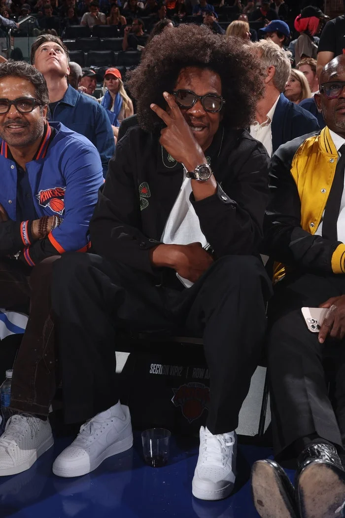A man sitting courtside at a sports event, wearing a casual outfit with sneakers, gestures with his hand near his face