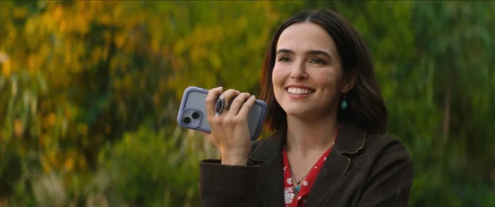 Person smiling, holding a phone, in an outdoor setting with greenery in the background