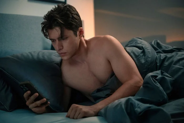 A shirtless man in bed looks intently at his phone, appearing focused and engaged