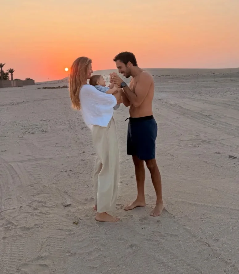 A couple stands on a beach at sunset, gazing lovingly at their baby, who they hold between them