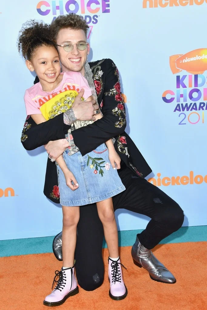 Person in floral jacket hugging a smiling child in a denim skirt on a red carpet at a Nickelodeon event