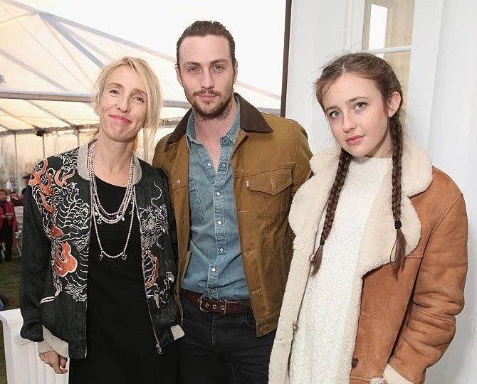 Three people standing together, smiling. The person on the left wears a patterned blazer, the middle person wears a casual jacket, and the third has a sherpa coat