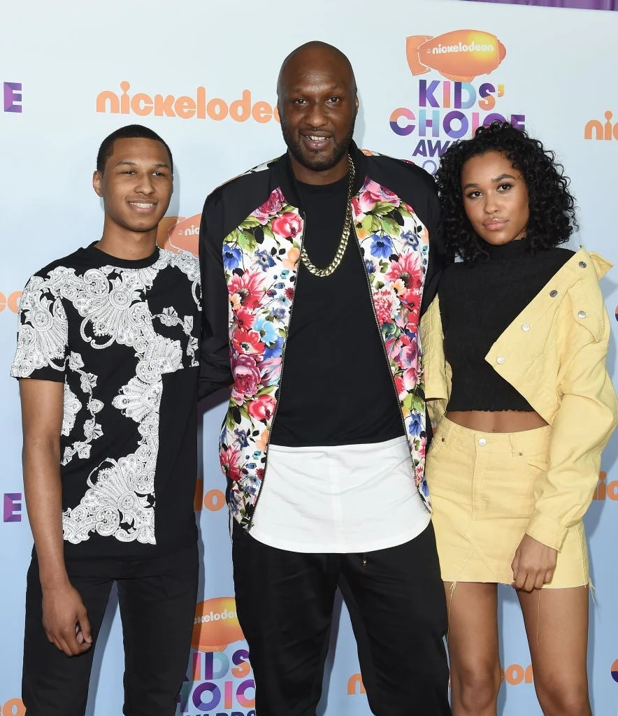 Three individuals pose at an event; the center person wears a floral jacket, while the others sport casual black-and-white and light-colored outfits