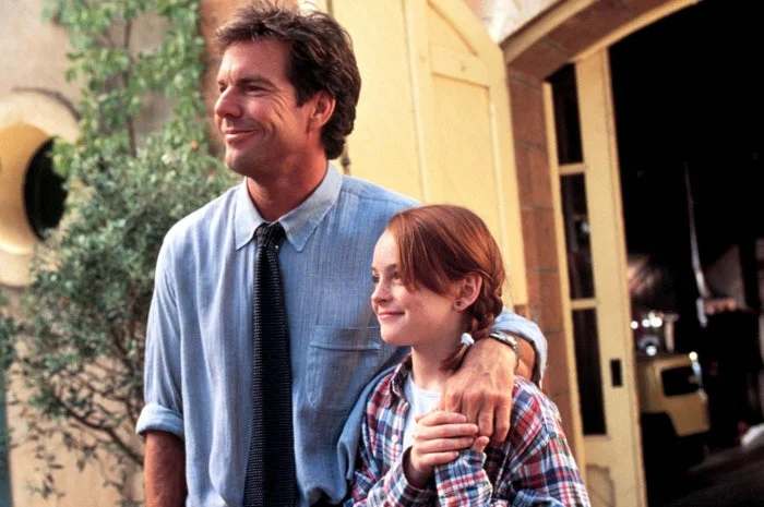 Two people standing outside a building. The man wears a collared shirt and tie; the young person wears a plaid shirt, smiling and looking ahead
