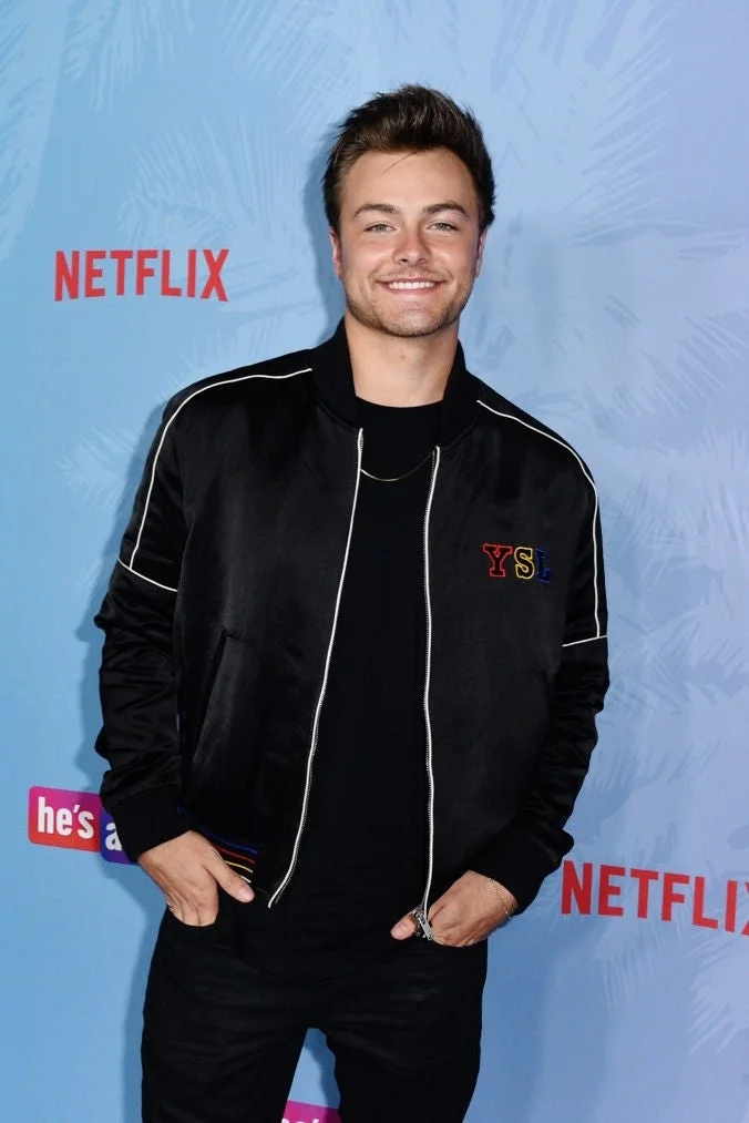 A person smiling at a Netflix event, wearing a casual black jacket over a t-shirt