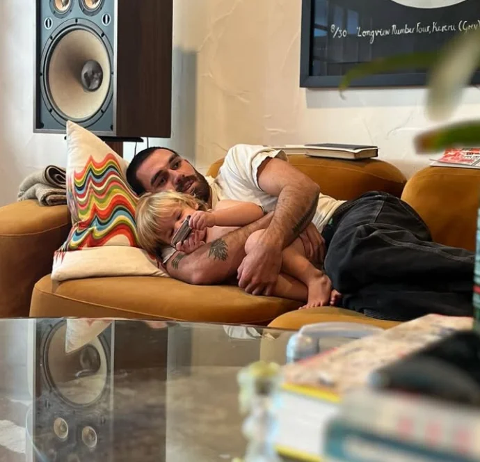 A man and a child cuddle on a sofa in a cozy living room, beside a colorful pillow and large speaker, creating a warm, relaxed scene