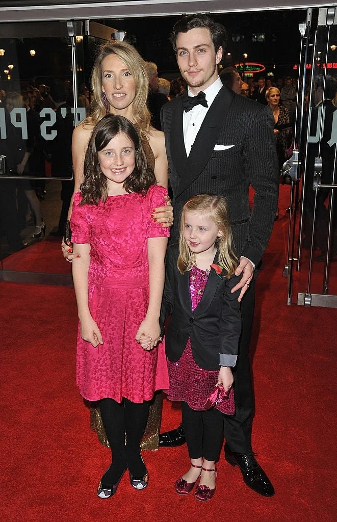 Two adults and two children pose on a red carpet. The adults are in formal attire, the man in a suit and the woman in a strapless dress