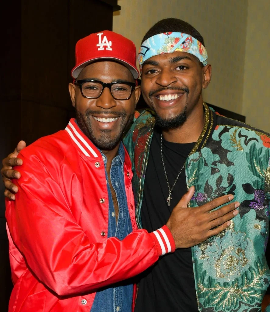 Two smiling individuals pose together. One wears a red jacket and cap; the other is in a floral jacket with a headband