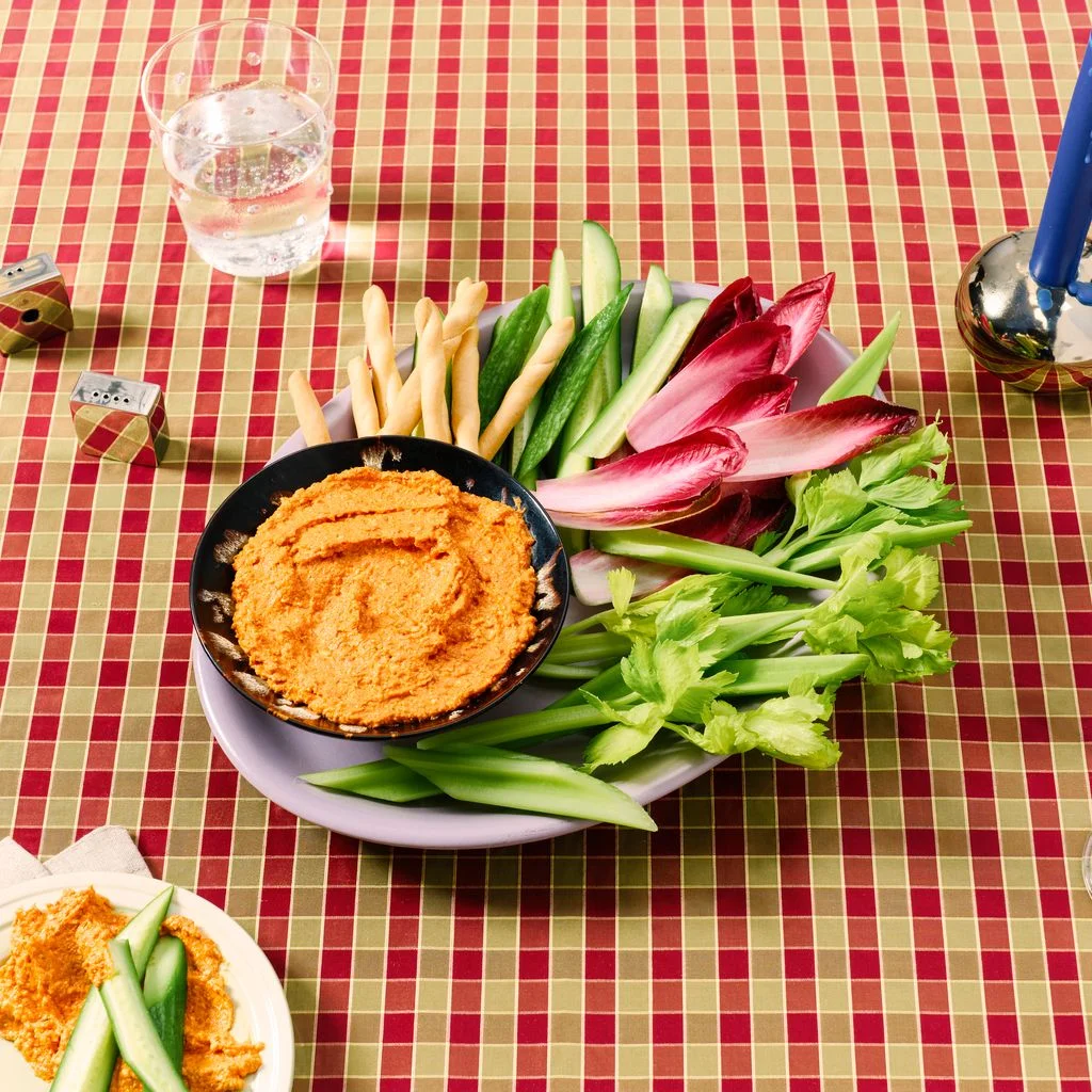 A creamy feta and sundried tomato dip with crudit on a checkered tablecloth with cocktails.
