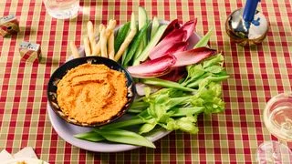 A creamy feta and sundried tomato dip with crudit on a checkered tablecloth with cocktails.