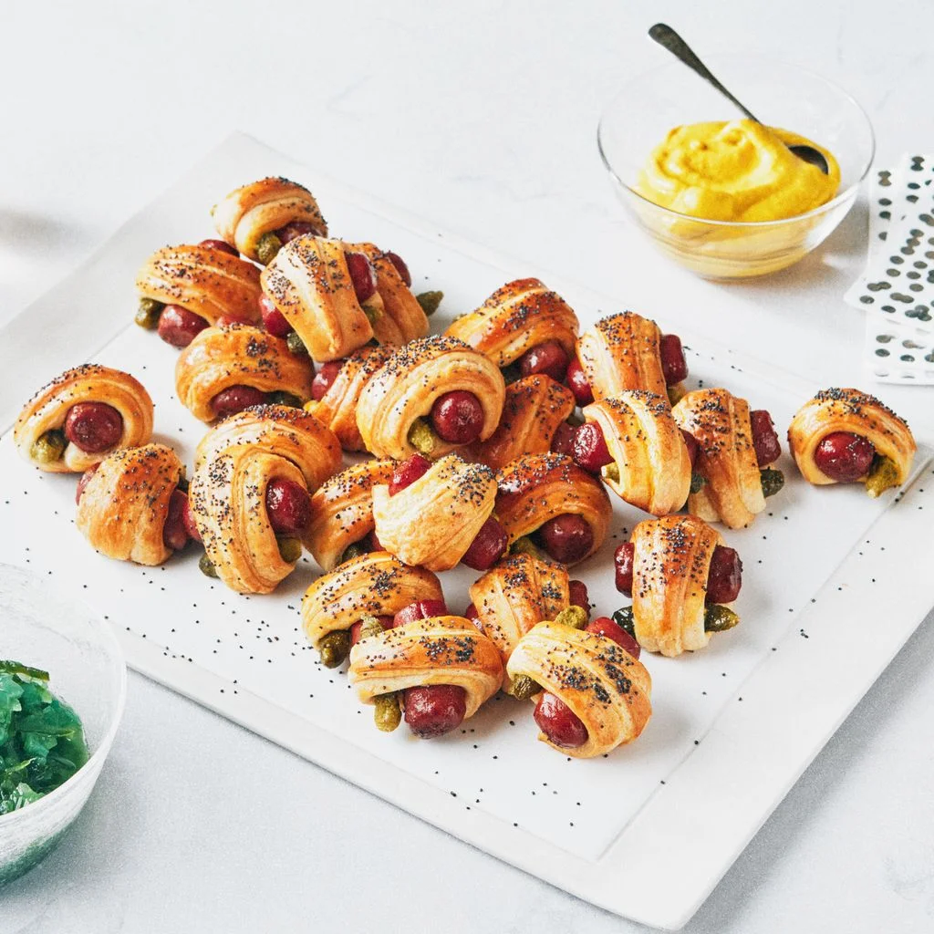 Pigs in a blanket topped with poppy seeds on an appetizer tray served with yellow mustard and Chicagostyle relish.