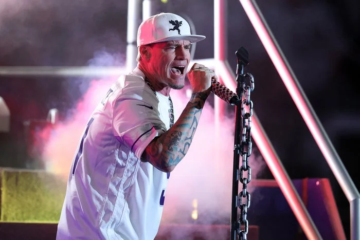 A performer wearing a cap and a white sports jersey sings energetically on stage, gripping the microphone with a tattooed arm