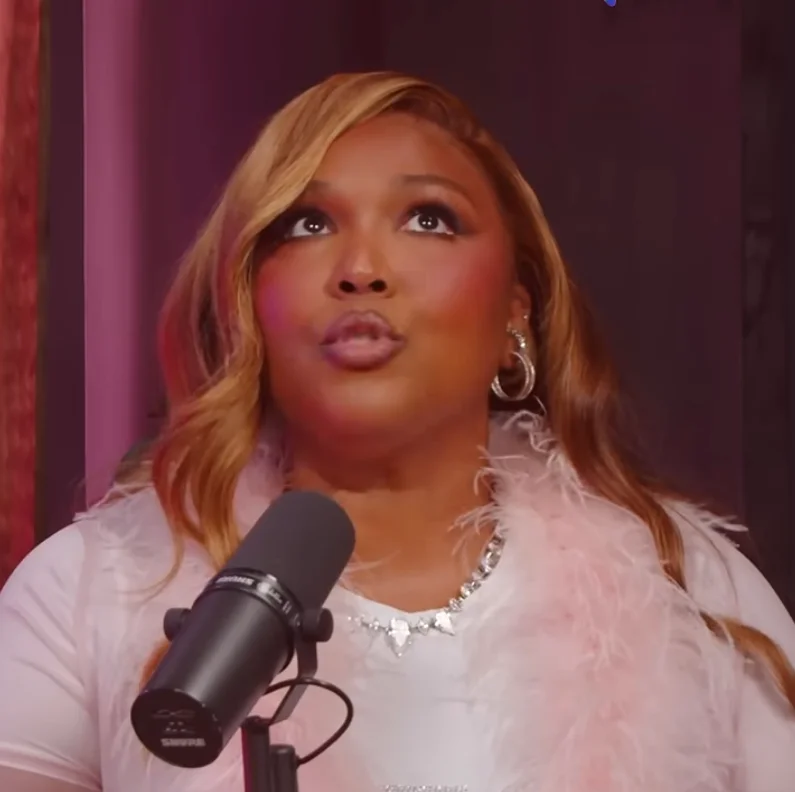 Lizzo with wavy hair speaks into a microphone, wearing a feathered accessory and earrings