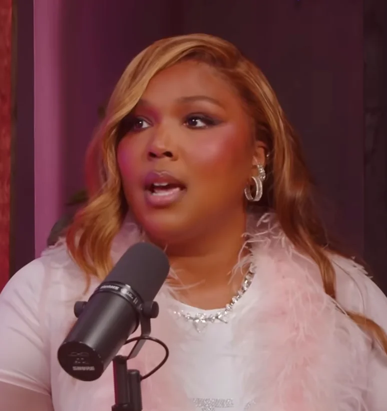Lizzo with wavy hair speaks into a microphone, wearing a feathered accessory and elegant jewelry
