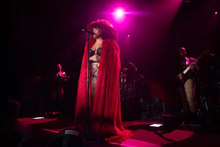 Lizzo performs on stage in an elaborate outfit, wearing a long, dramatic cape and a wide-brim hat, surrounded by band members