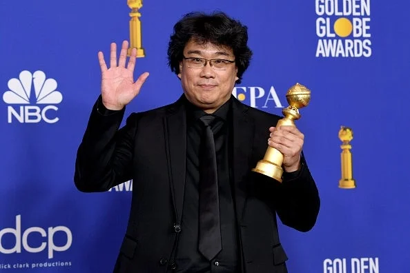 Bong Joon-Ho in a black suit holding a Golden Globe award, smiling, and waving at a Golden Globe Awards event