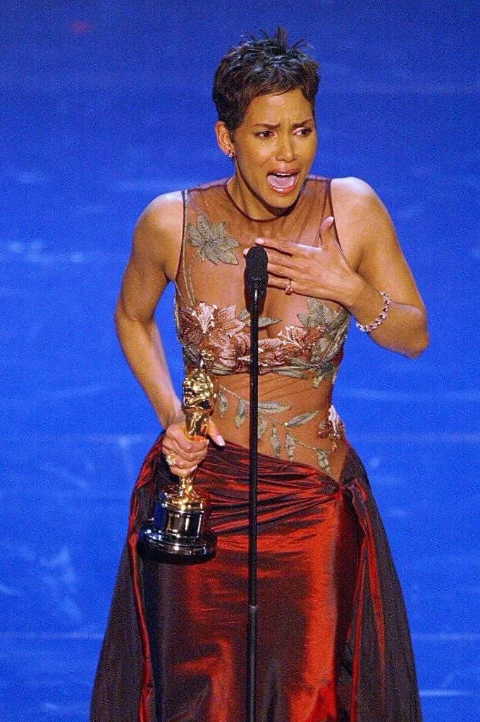 Halle Berry in an elegant gown, emotional, holding an award and speaking at a microphone on stage