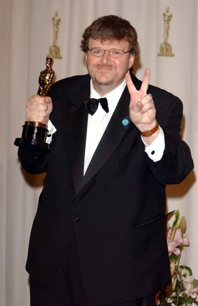 Michael Moore  in a tuxedo holds an Oscar statuette and makes a peace sign