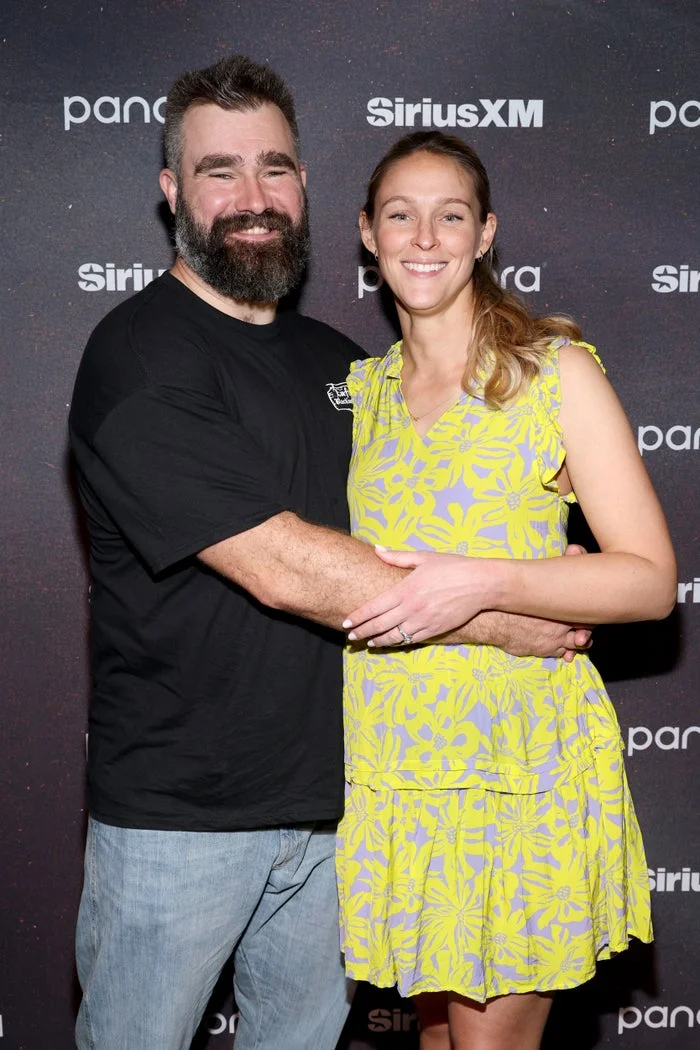 A couple poses together at a media event. The man wears a casual t-shirt and jeans; the woman wears a floral dress