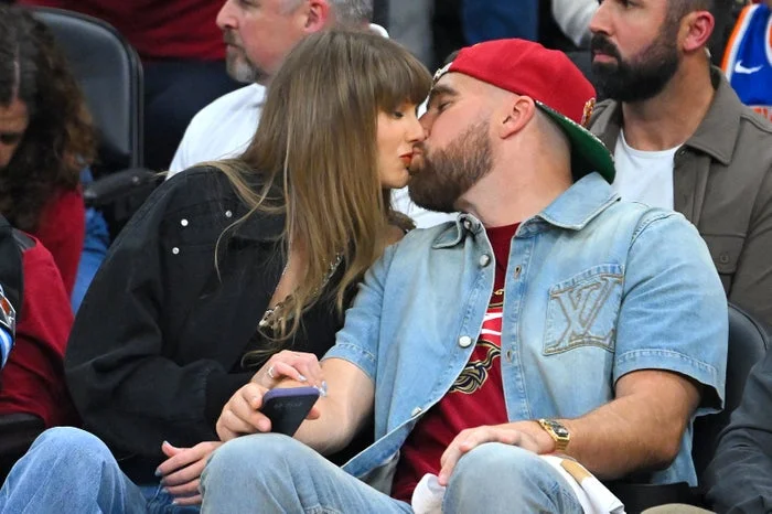 A couple sitting courtside at a sports event shares a kiss. The man wears a denim jacket and cap, while the woman has long hair and a casual outfit