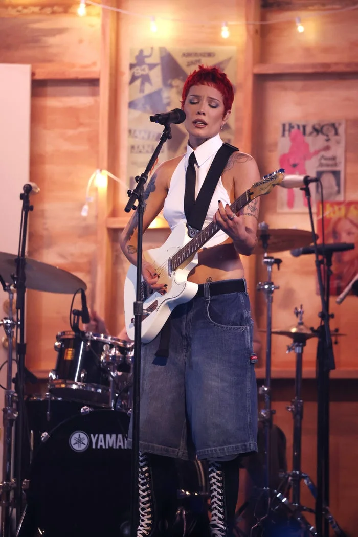 Halsey performing on stage with a guitar, wearing a sleeveless shirt, tie, denim shorts, and knee-high boots