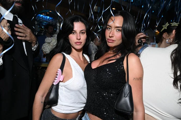 Two women posing at a party, one in a white tank top with pink hair clip on her bag, the other in a black sequined top. Balloons in background