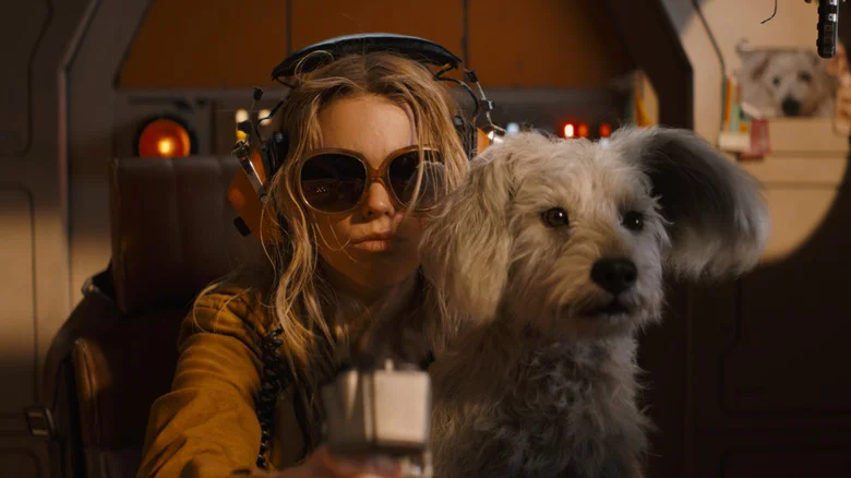 Milly Alcock as Supergirl wearing sunglasses and sitting at the controls of a spaceship with her dog Krypto on her lap in Supergirl