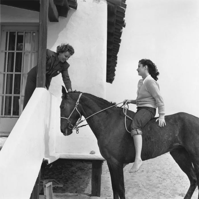 A young Elizabeth Taylor on horseback smiles at a woman leaning from a porch