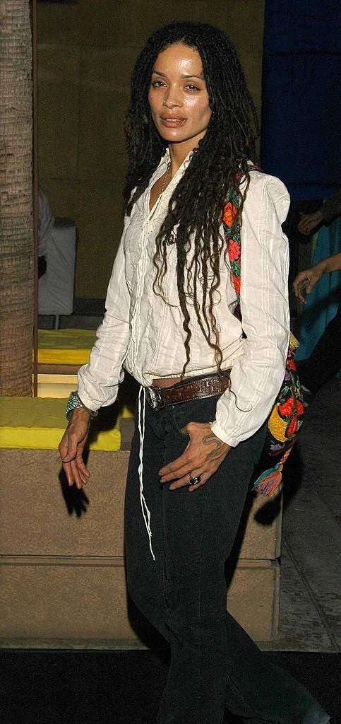 Person with long dreadlocks in a casual outfit featuring a white textured blouse and black pants, carrying a colorful patterned bag