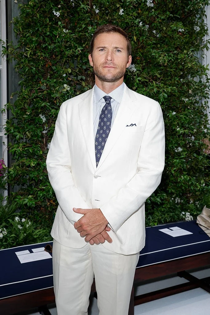 Person in a white suit with a patterned tie stands in front of greenery at an outdoor event