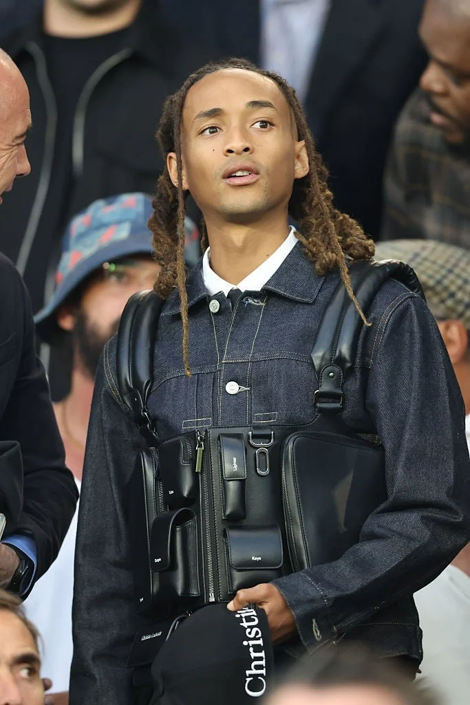 Person with long hair wears a denim jacket and a harness vest at a public event, looking up with a neutral expression